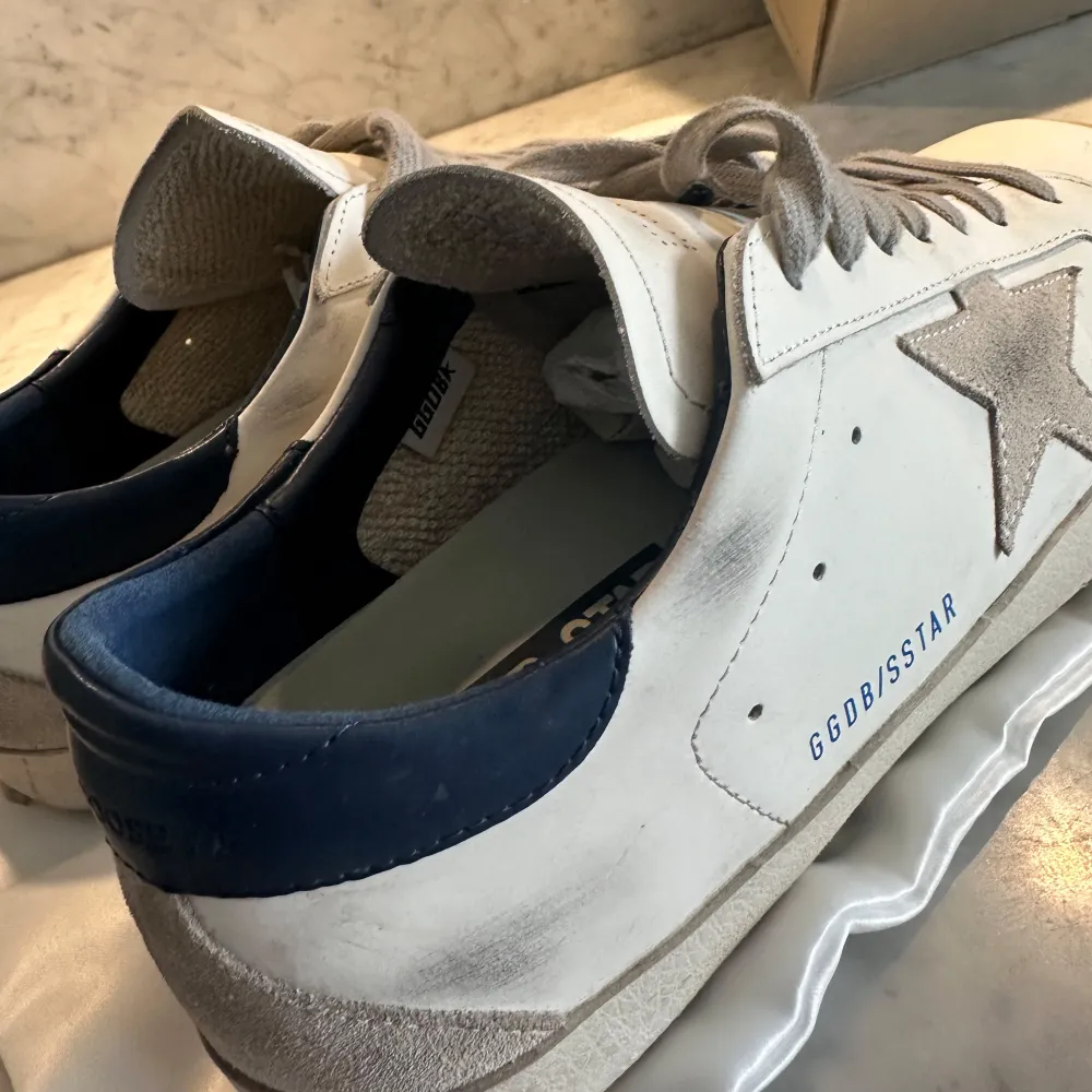 Snygga vita sneakers från Golden Goose med en ikonisk stjärndetalj på sidan. Skorna har en mörkblå häl och grå snörning. Jätte bra skick har bara en liten skada på baksidan av sulan. Bara att höra av er vid frågor. Box, påse och allt annat ingår. Är också öppen för byten.. Kengät.
