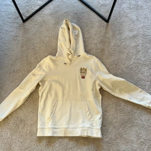 Vit/Creme hoodie från Morris - Säljer en stilren vit hoodie från Morris med broderad logga och brittisk flagga på bröstet. Tröjan har en klassisk känguruficka och justerbar huva med snören. Perfekt för en avslappnad stil.