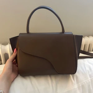 Brun handväska i skinn - Snygg brun handväska i skinn med ett stilrent och minimalistiskt utseende. Väskan har ett handtag och en asymmetrisk flik framtill. Perfekt för att bära dina dagliga nödvändigheter med stil.