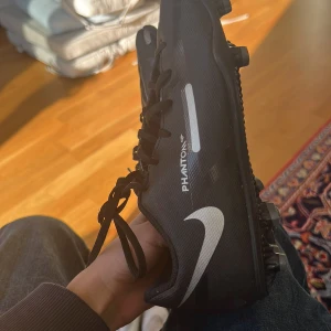 Svarta fotbollsskor från Nike - Säljer ett par svarta Nike Phantom fotbollsskor med vita detaljer. Skorna har en modern design med snörning och dobbar för optimalt grepp på planen. Perfekta för fotbollsträning och matcher.