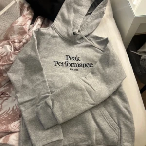 Grå hoodie från Peak Performance - Säljer en stilren grå hoodie från Peak Performance med logga på bröstet. Tröjan har en klassisk design med huva och snören samt ribbade muddar vid ärmslut och nederkant. Perfekt för en avslappnad look. Använd få gånger, inga defekter på.