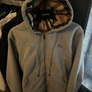 Grå hoodie från Burberry - Säljer en stilren grå hoodie från Burberry med klassiskt rutigt mönster i huvan. Tröjan har dragkedja framtill och praktiska fickor. Perfekt för en avslappnad look. Perfekt för alla årstider!