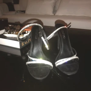 Svarta klackskor - Eleganta svarta klackskor med glittriga remmar som ger en lyxig känsla. Perfekta för en kväll ute eller en speciell tillställning. Skorna har en öppen tå och en smal klack som ger en stilren look.