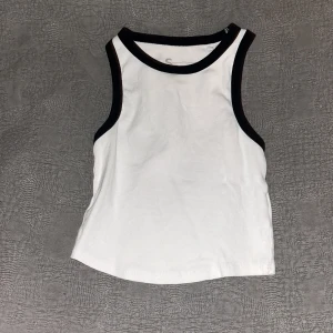 Svart vitt linne - Snygg vit ärmlös linnetopp med kontrasterande svarta kanter runt hals och ärmhål. Perfekt för varma dagar eller som lager under en jacka. Enkel och stilren design som passar till det mesta.