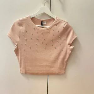  topp med pärlor - Säljer en söt rosa croppad topp från Bershka. Toppen har korta ärmar och är dekorerad med små pärlor framtill. Perfekt för en trendig look!