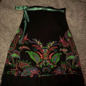 Svart kjol med färgglatt mönster - Snygg svart kjol med ett färgglatt, blommigt mönster i grönt, rött och blått. Kjolen har ett knytband i midjan för justerbar passform. Perfekt för att liva upp garderoben!