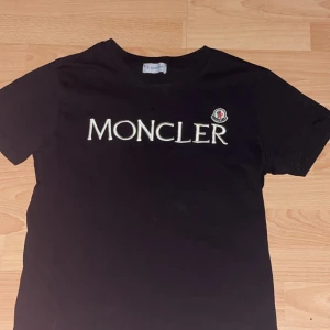 Svart moncler t-shirt  - Snygg svart t-shirt från Moncler med stort vitt logotryck på bröstet. Perfekt för en stilren look. S i storlek men passar även Medium 