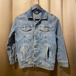 Ljusblå jeansjacka - Säljer en klassisk ljusblå jeansjacka med knappar framtill och två bröstfickor med lock. Jackan har en normal passform och långa ärmar. Perfekt för vår och sommar!