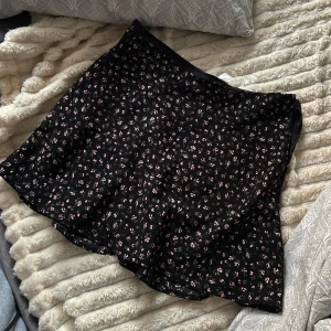 Svart blommig kjol från H&M Divided - Säljer en svart kortkjol från H&M Divided med ett sött blommönster i rosa och vitt. Kjolen har en normal passform och är perfekt för både vardag och speciella tillfällen. Den är tillverkad i ett lätt och bekvämt material.