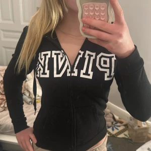 Svart huvtröja från Victoria's Secret PINK - Säljer en svart huvtröja från Victoria's Secret PINK med dragkedja och stor vit logga på framsidan. Tröjan har långa ärmar och en praktisk huva med snören. Perfekt för en avslappnad stil.