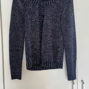 Mörkblå och silverglittrig stickad kofta från Juno - Säljer en snygg svart och vit stickad kofta från Juno. Koftan har långa ärmar och knappar framtill. Perfekt för kyliga dagar och ger en stilren look. Passar bra till både jeans och kjol.