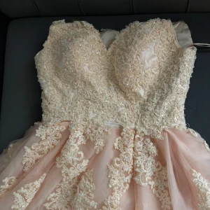 Champagnefärgad klänning med spetsdetaljer - Elegant champagnefärgad klänning med vacker spets och broderade detaljer. Axelbandslös design med en voluminös kjol som ger en romantisk känsla. Perfekt för speciella tillfällen.