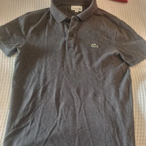 Grå pikétröja från Lacoste - Säljer en klassisk grå pikétröja från Lacoste med den ikoniska krokodilloggan på bröstet. Tröjan har korta ärmar och knappar vid kragen. Perfekt för en stilren och avslappnad look.