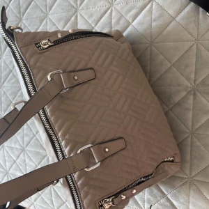 Grå grön don donna väska med dragkedjor - Snygg grå grön väska med quiltat mönster och flera dragkedjor. Väskan har både handtag och en justerbar axelrem i svart och beige. Perfekt för att bära med sig det viktigaste med stil.