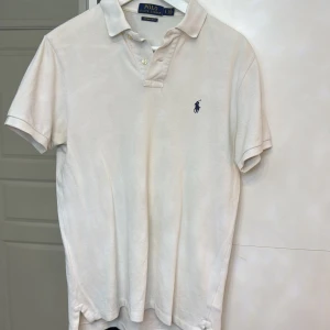 Vit pikétröja från Ralph Lauren - Säljer en klassisk vit pikétröja från Ralph Lauren med den ikoniska loggan broderad på bröstet. Tröjan har korta ärmar och en stilren krage med knappar. Perfekt för en avslappnad men stilfull look.