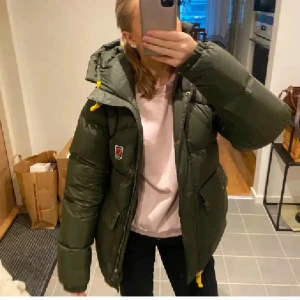 Grön dunjacka från Fjällräven - Säljer en grön dunjacka från Fjällräven, perfekt för kalla dagar. Jackan har en pufferdesign med huva och gula detaljer. Den är nästan helt som ny, köptes på naturkompaniet i mars