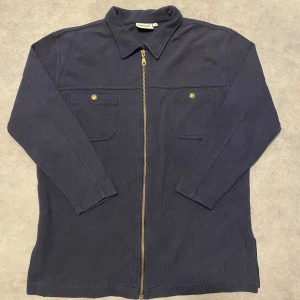 Mörkblå overshirt från Cappuccini - Säljer en stilren mörkblå overshirt från Cappuccini med dragkedja och två bröstfickor med knappar. Perfekt för lager-på-lager-stil. Den har långa ärmar och en klassisk krage. Skick 10/10