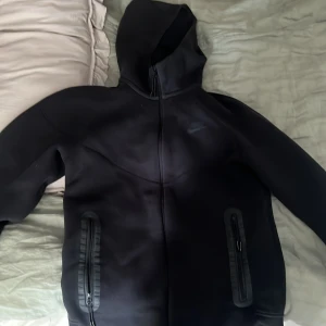 Nike tech kofta - Säljer en svart hoodie från Nike med dragkedja och huva. Jackan har långa ärmar och praktiska fickor med dragkedjor. Perfekt för en sportig look eller avslappnade dagar.