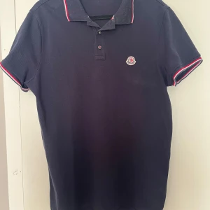 Mörkblå pikétröja med röd och vit detalj - Snygg mörkblå pikétröja från Moncler med klassisk krage och knappar. Tröjan har röda och vita detaljer på krage och ärmslut samt Monclers logga på bröstet. Perfekt för en stilren look.