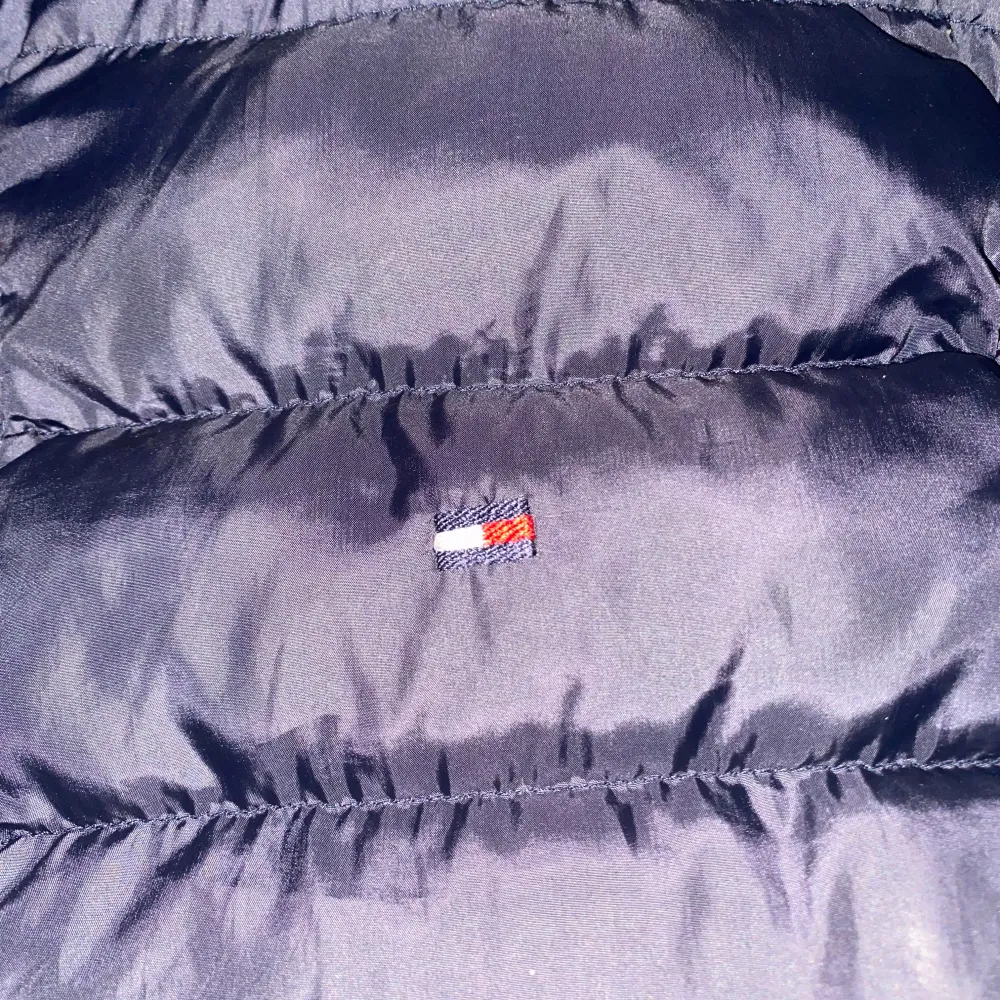 Snygg marinblå dunväst från Tommy Hilfiger med dragkedja och diskret logga på bröstet. Perfekt för lager-på-lager under kyliga dagar. Västen har praktiska fickor med dragkedja för säker förvaring.. Takit.