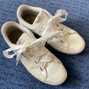 Vita sneakers från Puma - Snygga vita sneakers från Puma med breda satinliknande snören. Skorna har en rund tå och är perfekta för en stilren look. Tillverkade i syntetmaterial med en bekväm passform.