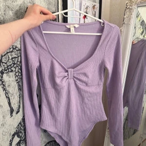 Ljuslila ribbad body med rosettdetalj - Säljer en ljuslila ribbad långärmad body med söt rosettdetalj framtill och v-ringning. Toppen är figurnära och har en mjuk, stretchig känsla. Perfekt att styla med jeans eller kjol för en trendig look. Jätte cute ger lite Rapunzel vibes!!🎀💜