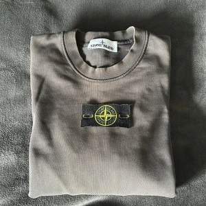 Grå sweatshirt från Stone Island - Säljer en grå sweatshirt från Stone Island med klassisk patch på armen. Tröjan har rund halsringning och är tillverkad i mjukt bomullsmaterial. Perfekt för en stilren och avslappnad look. Som man kan se på bilderna är tröjan lite solblekt och har en mycket liten skada där patchen ska sitta, som man inte ser när märket sitter på. Vid frågor tveka inte på att höra av dig!
