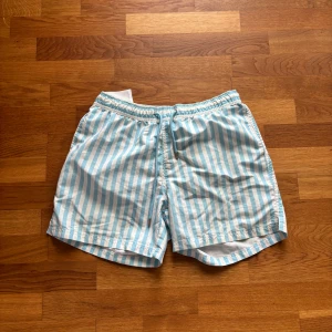 Blåvit randiga badshorts - Säljer ett par blå och vitrandiga badshorts med elastisk midja och snörning. Shortsen har en klassisk passform och är perfekta för sommardagar vid stranden eller poolen.