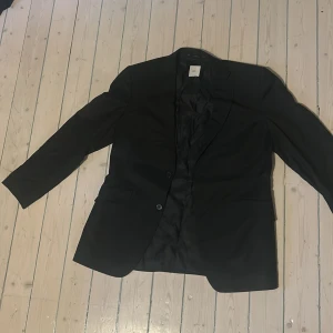 Svart kavaj från Bläck - Klassisk svart kavaj från Bläck med två knappar framtill och innerfoder. Kavajen har smal passform och enkla detaljer, perfekt för formella tillfällen. Fickor framtill och innerfickor. Stilren och tidlös design.