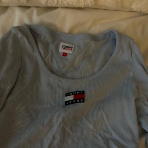 Ljusblå ribbad topp från Tommy Jeans - Säljer en ljusblå ribbad topp från Tommy Jeans med rund halsringning och broderad logga framtill. Perfekt för en avslappnad och stilren look.