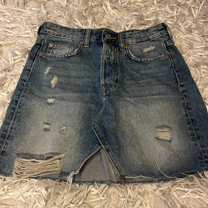 Jeanskjol med slitningar - Säljer en klassisk blå jeanskjol med slitna detaljer. Kjolen har fem fickor, knapp och dragkedja framtill samt en något A-linjeformad passform. Perfekt för en avslappnad och trendig look. Perfekt till sommaren.