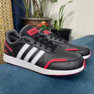 Svarta Adidas sneakers med röda och vita detaljer - Snygga svarta Adidas sneakers med klassiska vita ränder på sidorna och röda detaljer. Skorna har svart snörning, vit sula och röd undersida. Perfekta för dig som gillar sportig stil och vill sticka ut lite extra.