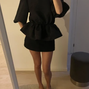 Svart blus med puffärmar  - Säljer en svart blus från NA-KD med markerad midja och volang. Blusen har puffiga trekvartsärmar och strukturmönster i tyget. Perfekt för dig som vill sticka ut med en trendig och elegant look.