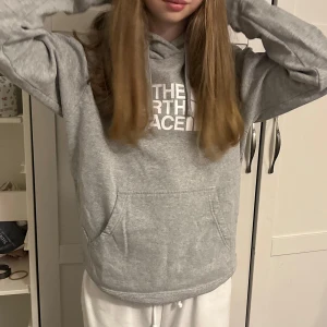 Grå hoodie från The North Face - Säljer en klassisk grå hoodie från The North Face med vit logga på bröstet. Tröjan har huva och magficka, perfekt för en avslappnad stil. Passar dig som gillar bekväma och stilrena plagg.