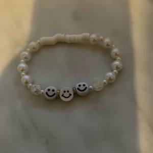 Pärlarmband med smileys - Snyggt armband med vita pärlor och tre glada smiley-ansikten framtill. Perfekt accessoar för att liva upp din outfit med lite extra glädje och personlighet.