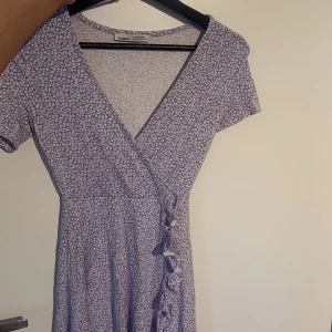 Ljuslila blommig omlottklänning från Pull&Bear - Supersöt ljuslila omlottklänning från Pull&Bear med små vita blommor. Klänningen har korta ärmar, v-ringning och knyt i sidan. Perfekt för vår och sommar! (Tyvärr aldrig använd då den är lite kort i min smak, men sååå fin och somrig) 
