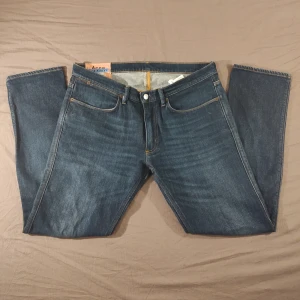 Acne Studios Blå Konst Jeans - Mörkblå jeans från Acne Studios Blå Konst med klassisk femficksdesign och slimade ben. Jeansen har orangea sömmar och en ikonisk acne patch bak i midjan. Perfekta för dig som gillar stilrena och tidlösa jeans.