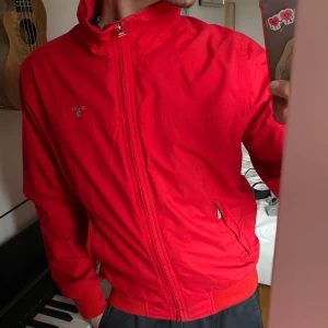 Röd vindjacka från GANT - Säljer en röd vindjacka från GANT i klassisk modell. Jackan har dragkedja framtill, ståkrage och två fickor med dragkedja. Perfekt för blåsiga dagar och enkel att matcha med olika outfits. Säljer då jag inte använder. Den är köpt på second hand och den flagnar lite på insidan, men köpte den ändå för den är fin på utsidan. Säljer nu för bra pris
