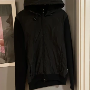 Svart jacka med huva från Porsche x BOSS - Säljer en svart jacka från Porsche x BOSS med huva och dragkedja framtill. Jackan har en stilren design med släta paneler och ribbade muddar. Perfekt för dig som gillar exklusiva samarbeten och snygg streetstil.