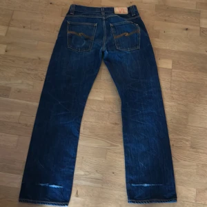Mörkblå jeansbyxor från Nudie Jeans - Säljer ett par mörkblå jeans från Nudie Jeans med klassisk femficksdesign och kontrastsömmar på bakfickorna. Byxorna har raka ben och normal passform, perfekt för en avslappnad stil.