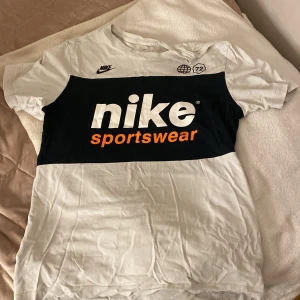Nike Sportswear t-shirt med tryck - Säljer en vit och svart Nike Sportswear t-shirt med stort tryck framtill i vitt och orange. Klassisk passform med korta ärmar och rund hals. Perfekt för en sommar dag.
