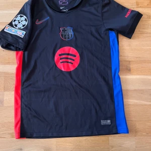 FC Barcelona svart fotbollströja Nike - Snygg svart FC Barcelona fotbollströja med röda detaljer och tryck, inklusive Spotify-logga på framsidan och Raphinha 11 på ryggen. Blå och röda paneler på sidorna, officiella märken på ärmen. Perfekt för fans!