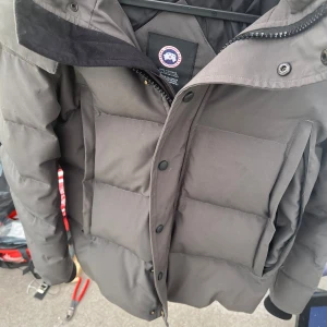 Svart dunjacka från Canada Goose med pälskrage - Säljer en svart dunjacka från Canada Goose med stor huva och avtagbar pälskant. Jackan har stora fickor framtill, dragkedja och knappar. Perfekt för kalla dagar och riktigt varm.