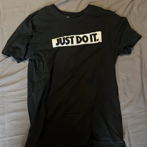 Svart t-shirt med 'JUST DO IT.' tryck från Nike - Svart t-shirt från Nike med det klassiska 'JUST DO IT.' trycket i vitt på bröstet. T-shirten har rund halsringning och korta ärmar. Perfekt för en sportig och avslappnad stil.