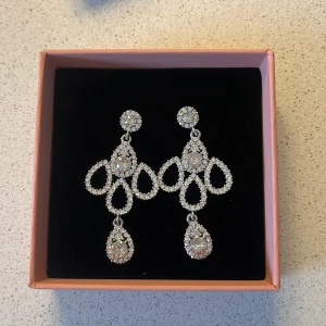 Glittriga silverfärgade örhängen - Säljer ett par stora, glittriga örhängen i silverfärg med gnistrande stenar från Caroline Svedbom som inte säljs längre! Örhängena har en elegant, droppformad design och passar perfekt när du vill sticka ut lite extra. Örhängena är vattentäta och är helt oanvända!