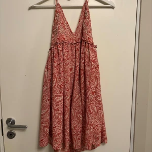 Röd- och vitmönstrad klänning från ZARA - Supersöt röd- och vitmönstrad klänning från ZARA med djup v-ringning och tunna axelband som knyts i nacken. Lätt och luftig modell med paisleymönster, perfekt för varma dagar. Kjolen är vid och faller fint.