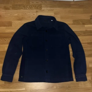 Mörkblå overshirt från d'once de la sense - Säljer en stilren mörkblå overshirt från d'once de la sense. Overshirten har två bröstfickor, klassisk krage och knäppning framtill. Perfekt att bära som lager över en t-shirt. Materialet känns mjukt och lite tjockare. Dm gärna vid frågor.