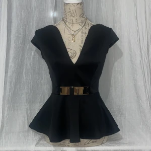 Svart peplum topp med bälte - Snygg svart peplum topp med djup v-ringning och korta ärmar. Toppen har ett brett, glansigt bälte i midjan som ger en markerad siluett. Perfekt för att skapa en elegant look.