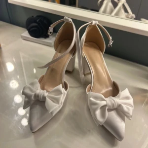 Vita pumps med rosett och ankelrem - Hej hej, säljer mina nya gulliga klackskor/pumps med en rosett på. Endast använda 1 gång. Super lätta att gå i på grund utav den breda klacken. Fina till bal, student, fest osv. Pris kan diskuteras 