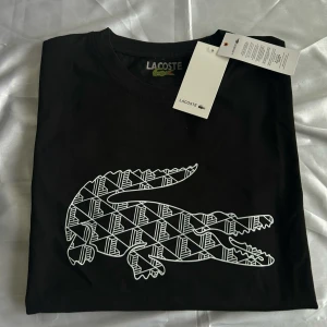 Svart t-shirt från Lacoste med grafisk krokodil - Snygg svart t-shirt från Lacoste med stort grafiskt krokodilmotiv i vitt på bröstet. Klassisk rund halsringning och korta ärmar. Perfekt för en stilren och sportig look.